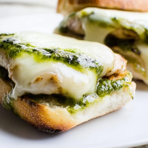 Golden-brown Pesto Chicken Melt halves resting on a wooden cutting board, revealing layers of gooey mozzarella and vibrant basil pesto. 